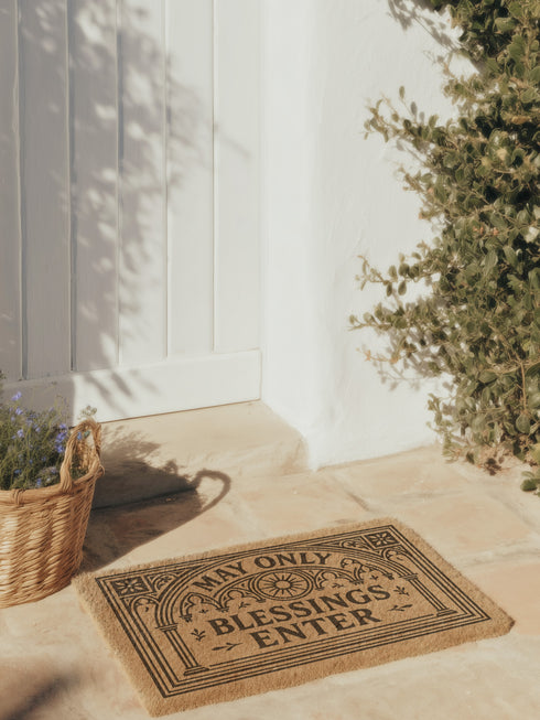 A rectangular natural brown coir doormat with a rugged texture and a durable black vinyl backing. The doormat features an intricate black design framed by a gothic arch with stylized quatrefoil details in the corners. Inside the arch, the words "MAY ONLY BLESSINGS ENTER" are printed in an elegant, bold serif font. A central sunburst medallion with radiating lines sits above the text, reinforcing the spiritual and protective theme of this gothic entryway welcome mat.